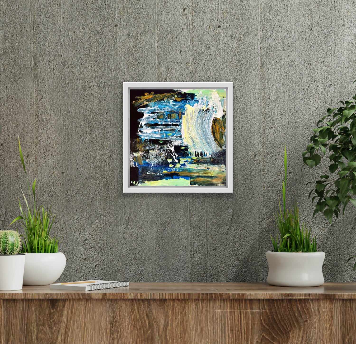 An abstract landscape painting of a river sits in a white frame in an elegant setting. It hangs on a grey textured concrete wall surrounded by green plants in white containers. There is a textured wooden sideboard underneath and the edge of a ficus is visible to the right.